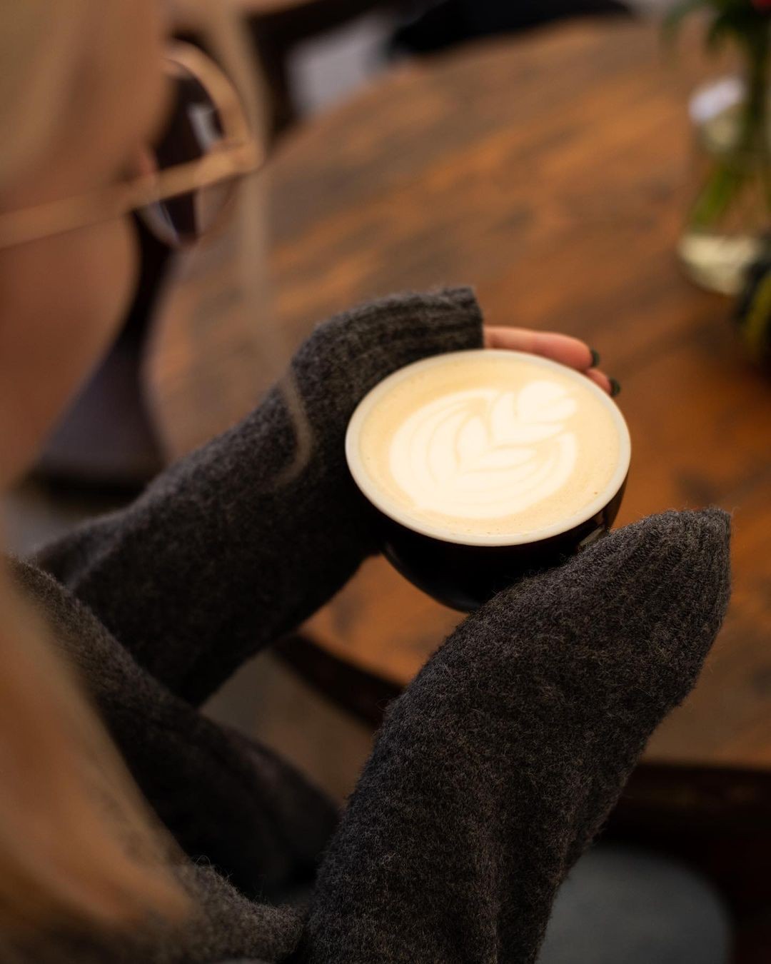 A cappuccino with heart-shaped latte art held in both hands over a wooden table at Drunken Coffee
